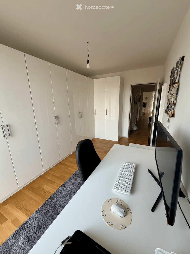 A modern, minimalist room with a white desk, black chair, keyboard, mouse, and screen. The room features light wooden flooring, white built-in wardrobes, and a grey rug.