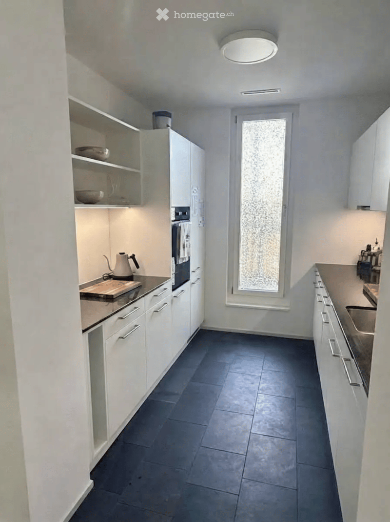 Modern kitchen with white cabinets, dark tiled floor, and stainless steel appliances. Features an oven, sink, and teapot on the counter.