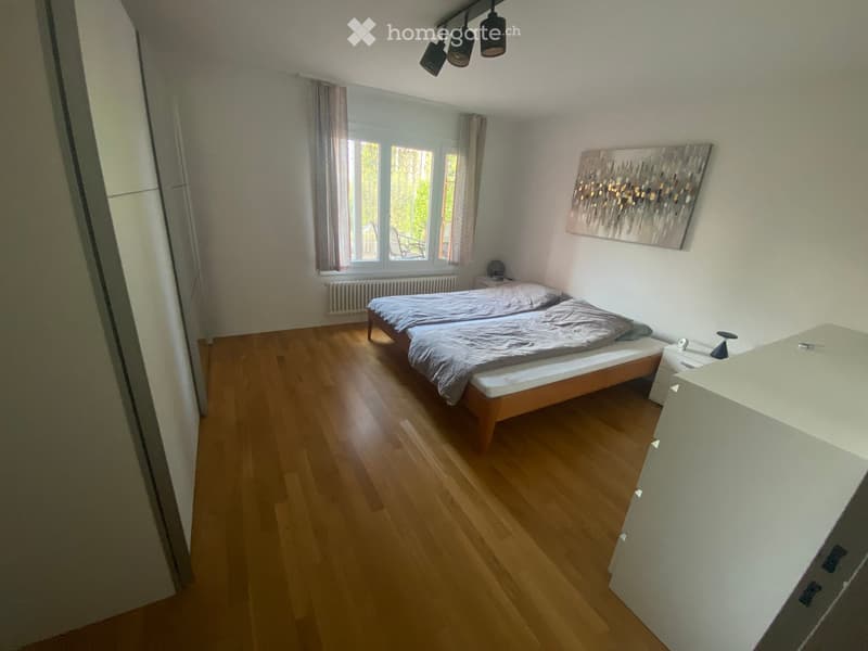 Cozy bedroom with wooden flooring, double bed, nightstand, painting on wall, and window with curtains.