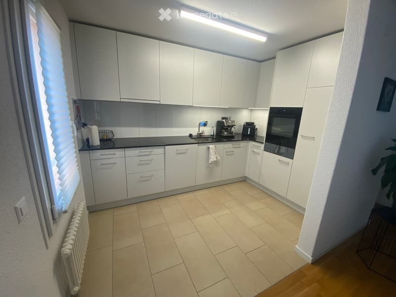 Modern kitchen with white cabinets, black countertops, and stainless steel appliances including a coffee maker and oven. Features a tiled floor and a window with blinds.
