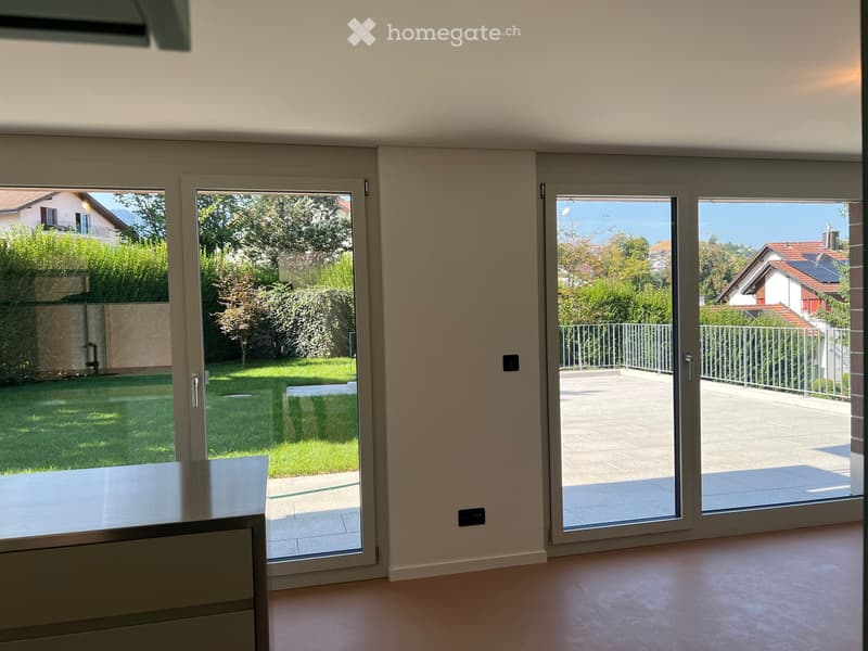 Bright, empty room with large sliding glass doors leading to a patio and garden. Light-colored walls and flooring.