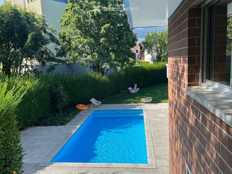 Private garden with a rectangular swimming pool, surrounded by neatly trimmed hedges, trees, and a lounge chair with inflatable tubes nearby.