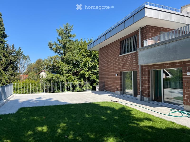 Modern two-story house with red brick facade, large windows, and a spacious balcony overlooking a well-maintained lawn and garden area.
