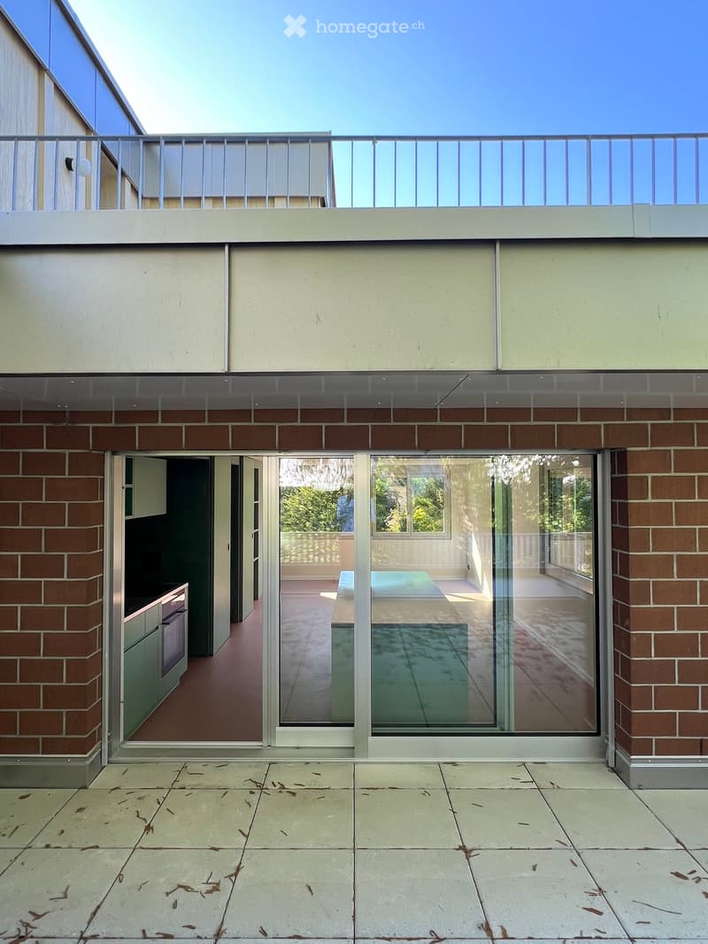 Modern building exterior with brick lower level, glass upper level, and a spacious balcony area with a view of greenery.