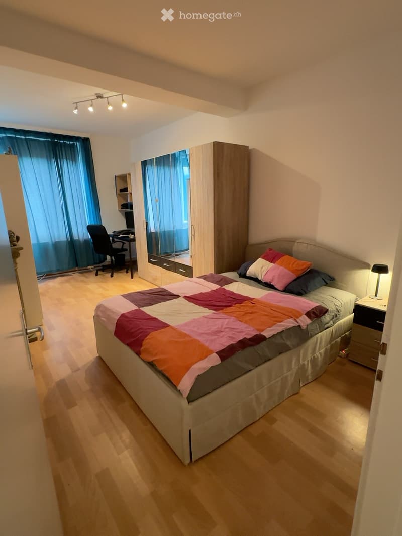 A well-lit bedroom with a wooden floor, a large bed with a colorful quilt, a nightstand with a lamp, a desk with a chair, and a wardrobe with a mirror.