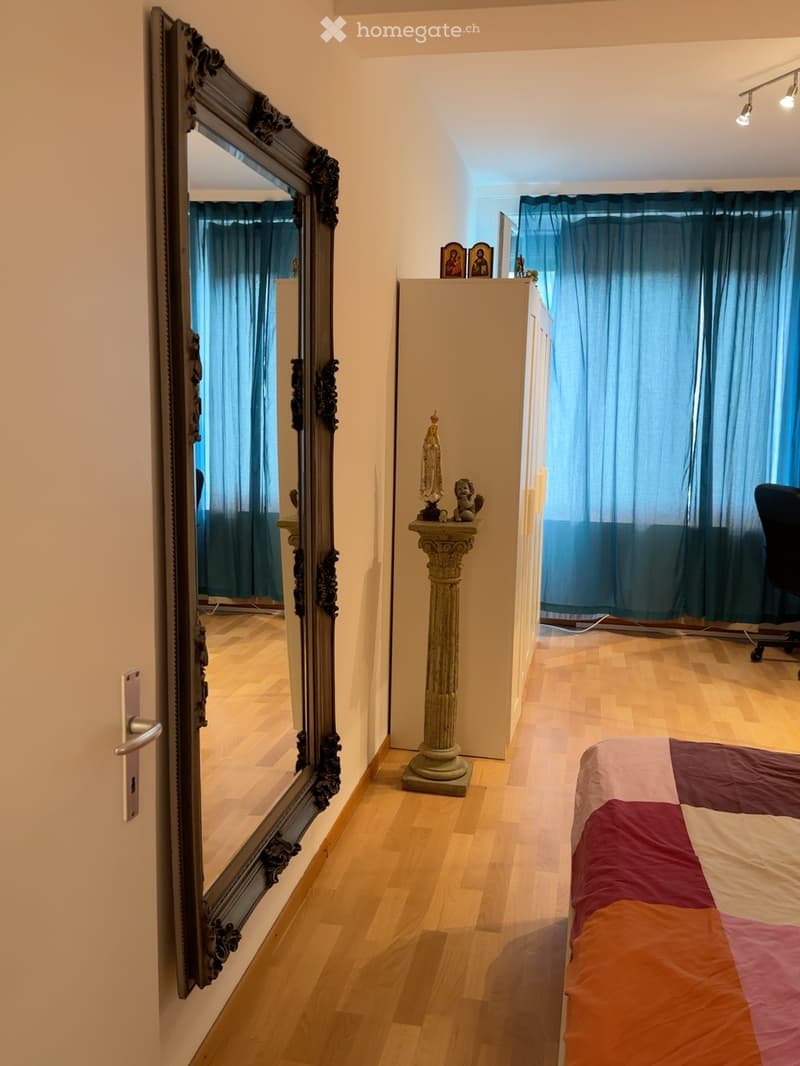 A well-lit bedroom with wooden flooring, a large ornate mirror on the wall, a bed with a colorful patchwork quilt, a small table with a statue, and a chair near the window with blue curtains.