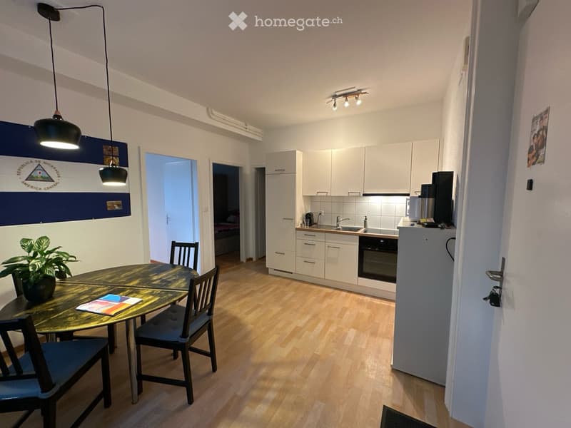 A modern, well-lit living space with a dining area and an open kitchen. The dining area features a round wooden table with four chairs, a potted plant, and some magazines. The kitchen has white cabinets, a sink, an oven, and a refrigerator. A screen is pl