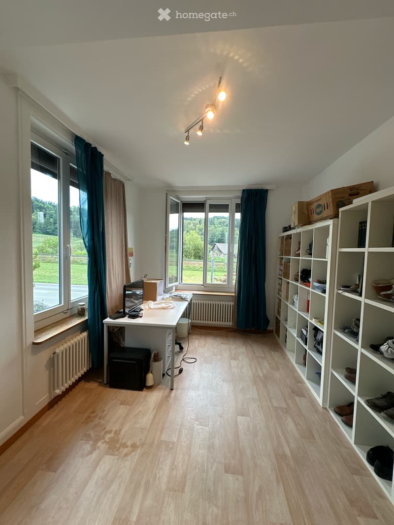 Bright room with wooden flooring, large windows with dark curtains, a desk with a laptop, a shelf with various items, and radiators under the windows.