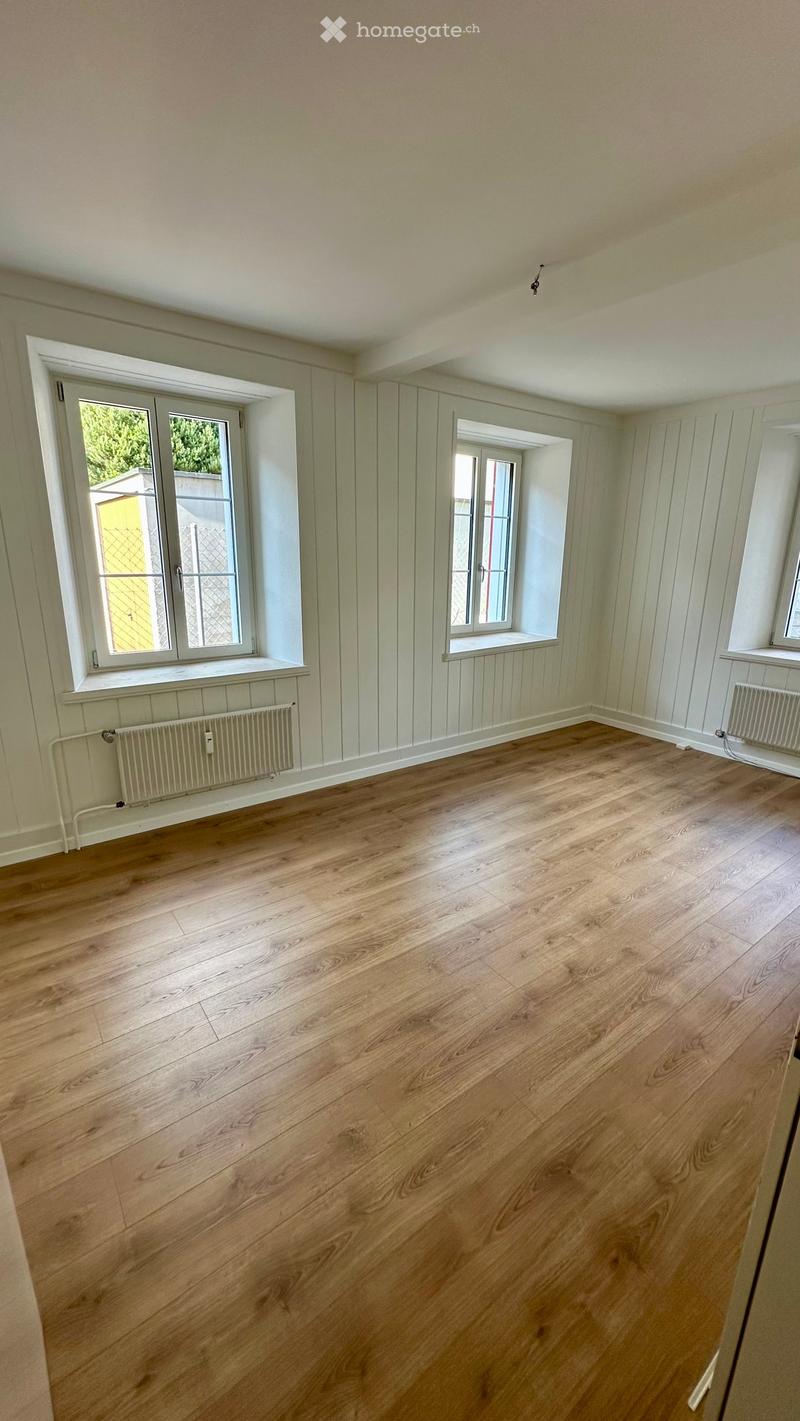Empty room with wooden flooring, white paneled walls, and multiple windows with white frames. Radiators are visible along the walls.