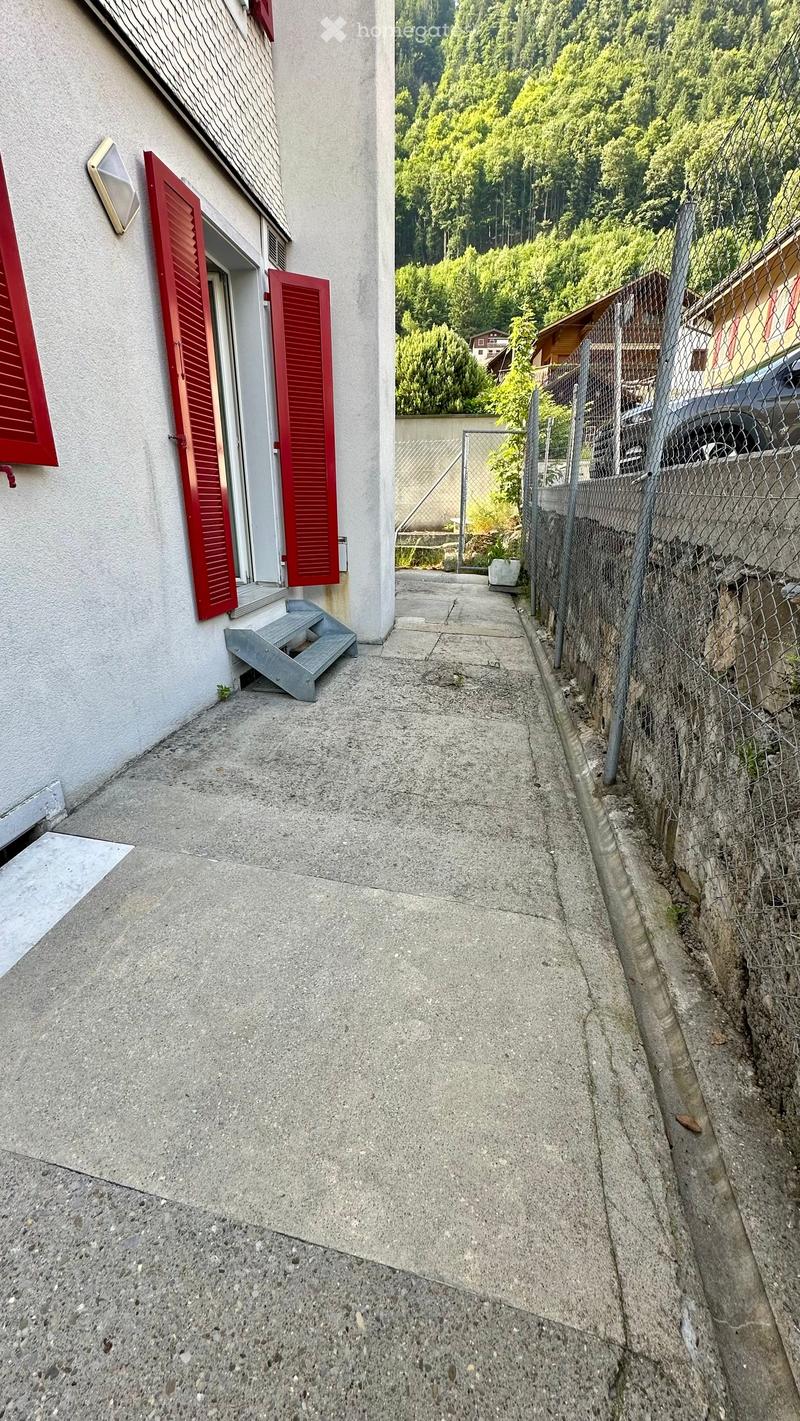 Narrow concrete pathway next to a building with red shutters and a small staircase leading to an entrance.