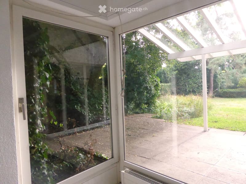 A covered balcony with large glass doors, white wooden frame, and a view of a green garden and lawn.