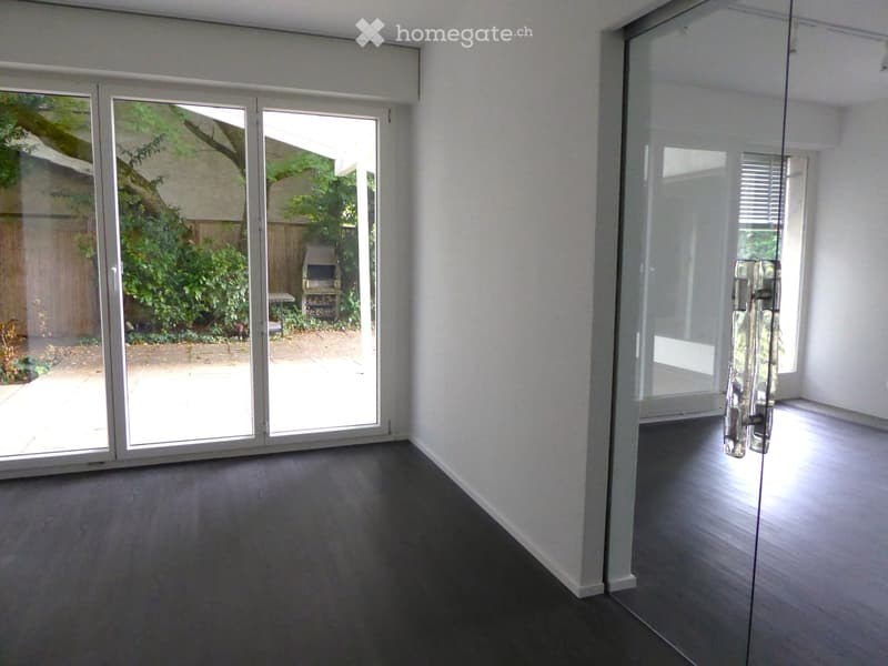Empty room with dark flooring, white walls, and large glass sliding doors leading to an outdoor area with greenery and a barbecue.
