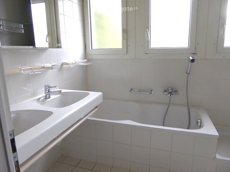 A clean, modern bathroom with white tiles, a double sink, a bathtub, and a large mirror above the sink.