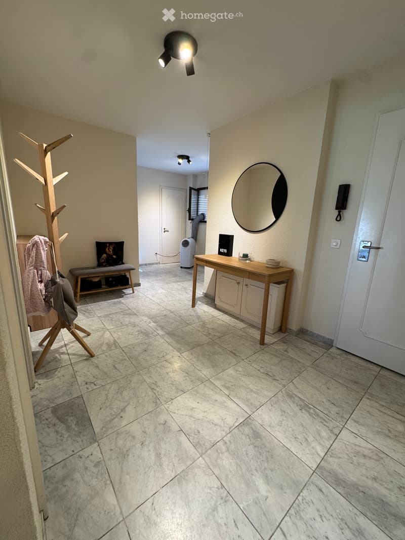 Clean, well-lit hallway with marble flooring, a round mirror, a small wooden table with a few items, a coat rack, and a bench with shoes and a bag.