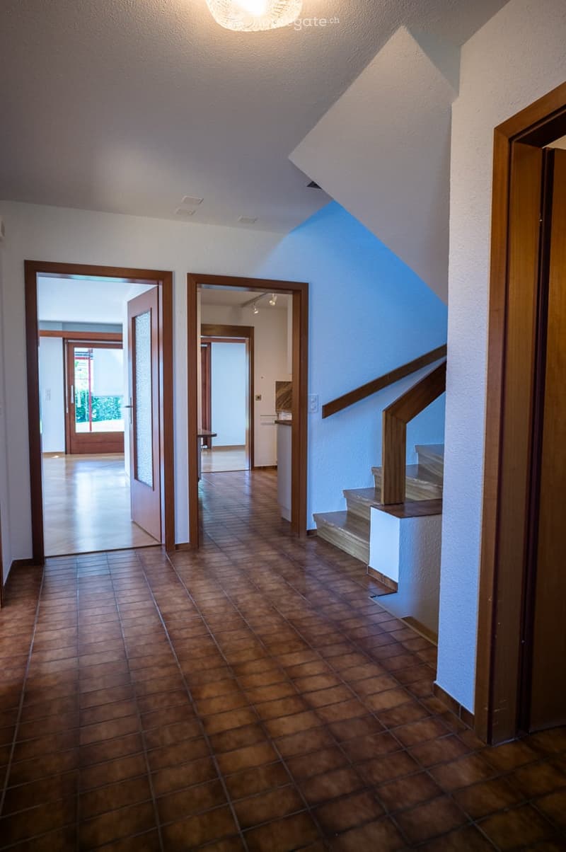 Spacious hallway with brown tiled flooring, white walls, and wooden doors leading to other rooms. A staircase with wooden railings ascends to an upper level.