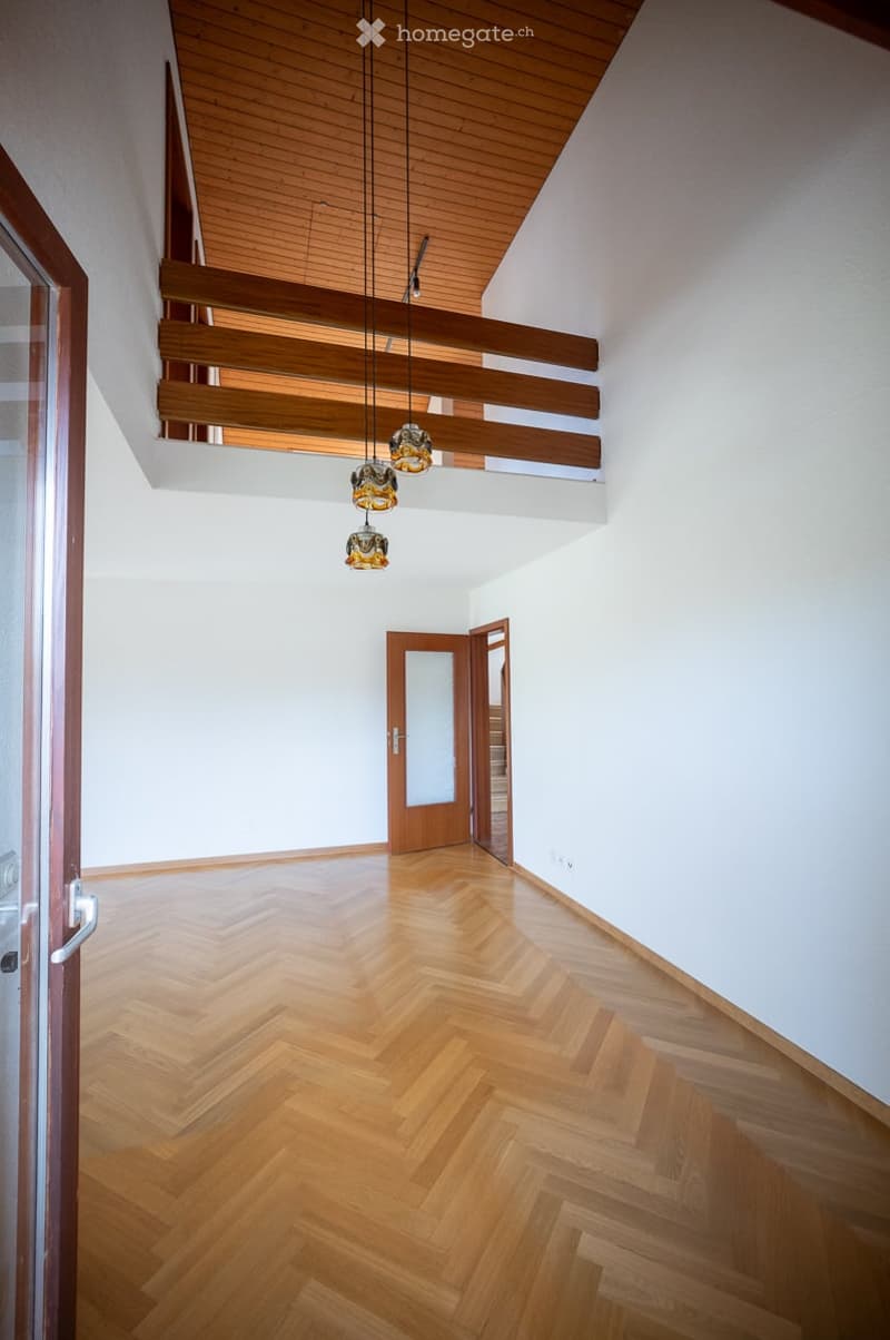 Empty hallway with wooden herringbone flooring, white walls, and a wooden ceiling with exposed beams. Features pendant lights and a wooden door leading to another room.