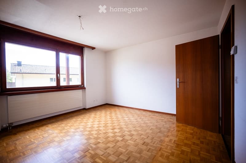 Empty room with wooden flooring, white walls, large window with wooden frame, and a closed wooden door.