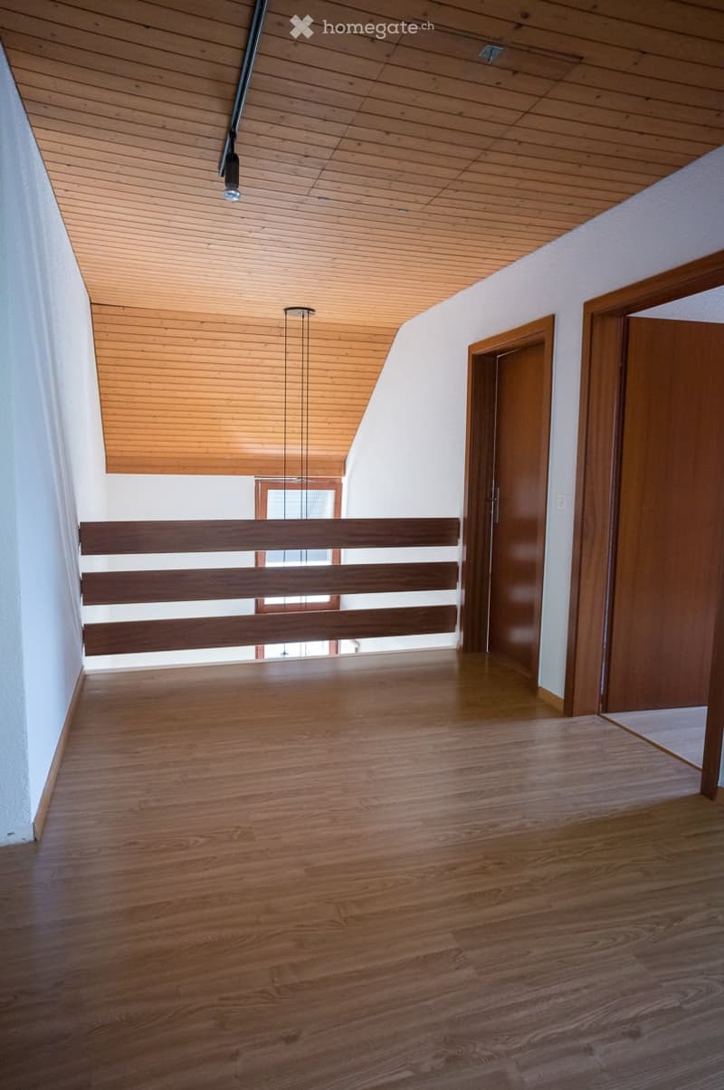 Empty hallway with wooden flooring, white walls, wooden ceiling, and a wooden railing leading to another room.