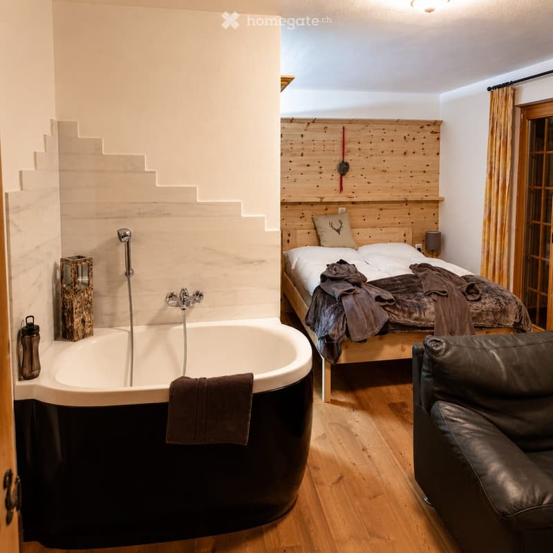 A modern, well-furnished room with a bathtub, a double bed, a leather sofa, and a decorative wooden wall. The room features wooden flooring, a large window with curtains, and a stylish lamp on the bedside table.