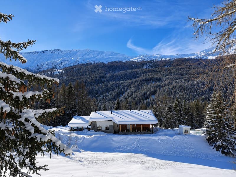 A snow-covered wooden house with a stone base, surrounded by snow-covered trees and mountains in the background.