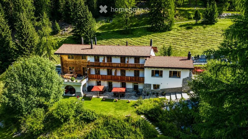 A two-story house with a wooden balcony, surrounded by a lush green lawn and trees. The house features a tiled roof, multiple windows, and outdoor seating areas with umbrellas.