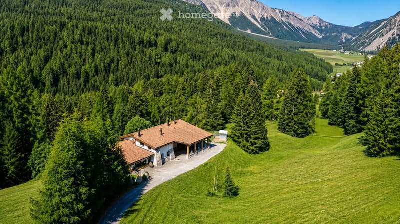 A two-story house with a red-tiled roof, surrounded by a lush green forest and mountains in the background.