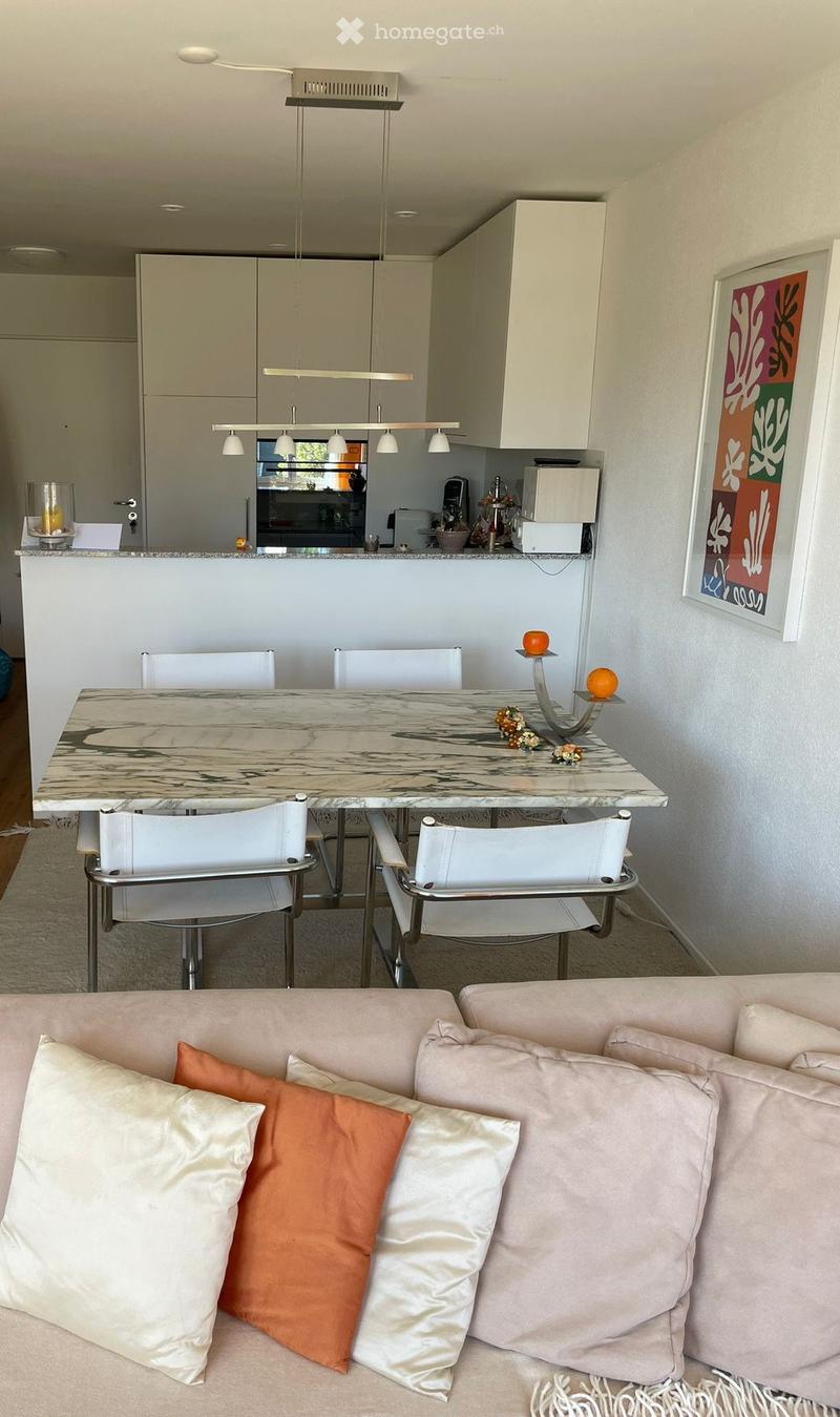 Modern open-plan living and dining area with a light-colored sofa, decorative pillows, a marble dining table with white chairs, and a kitchen area with cabinets and appliances.