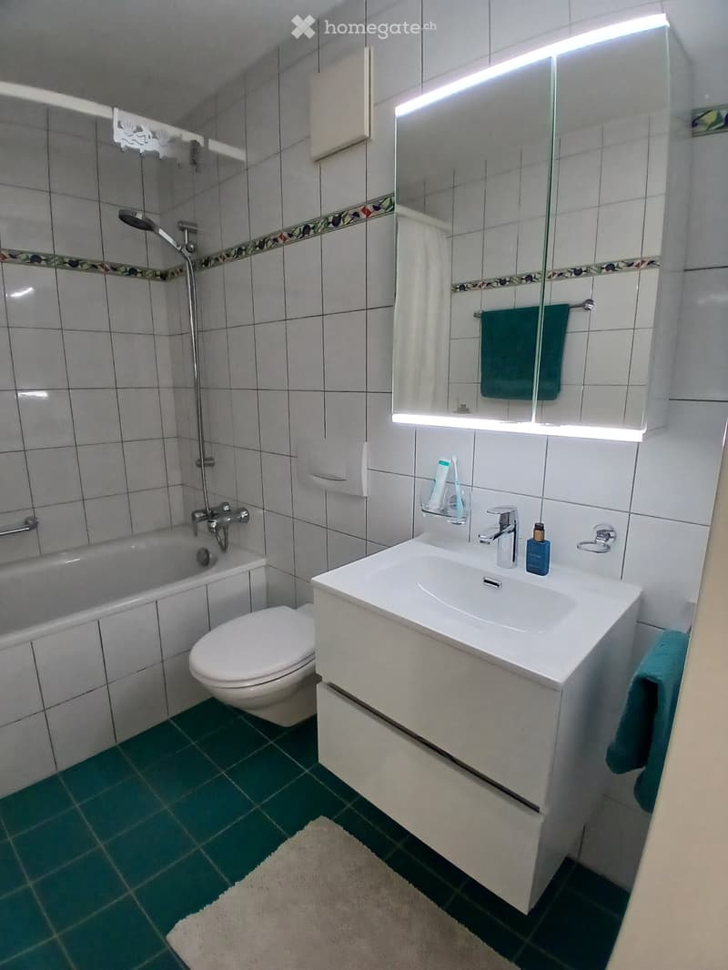 Modern bathroom with white tiles, green floor tiles, a white sink with a chrome faucet, a toilet, a bathtub with a shower, a large mirror with built-in lighting, and a towel hanging on the side.