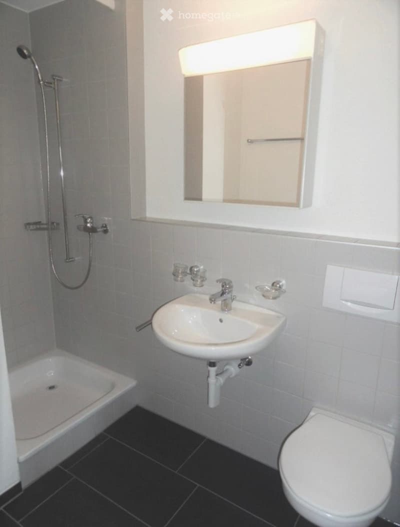 Modern bathroom with white tiles, a bathtub, a sink with a mirror, and a toilet. The showerhead is mounted on the wall.