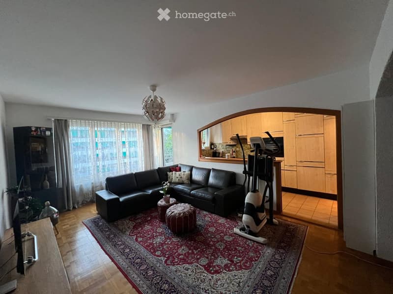 A modern living room with a black leather sectional sofa, two ottomans, a round stool, a patterned rug, an exercise bike, a large window with sheer curtains, and a kitchen area visible in the background.