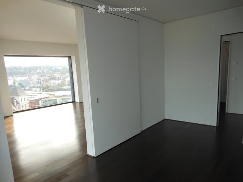 Empty room with wooden flooring, large window offering a city view, and white walls.