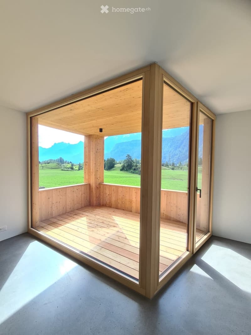 Open wooden balcony with large glass doors, overlooking a green field and mountains.