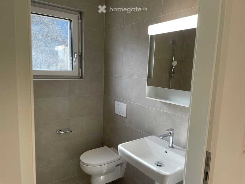 Modern bathroom with beige tiled walls, a white toilet, a white sink with a chrome faucet, a large mirror with built-in lighting, and a window providing natural light.