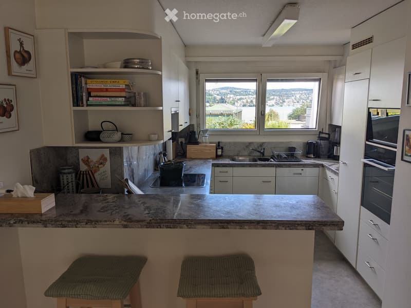 Modern kitchen with white cabinets, granite countertops, and a large window offering a scenic view. Includes appliances like an oven, dishwasher, and microwave, along with stools at the island.