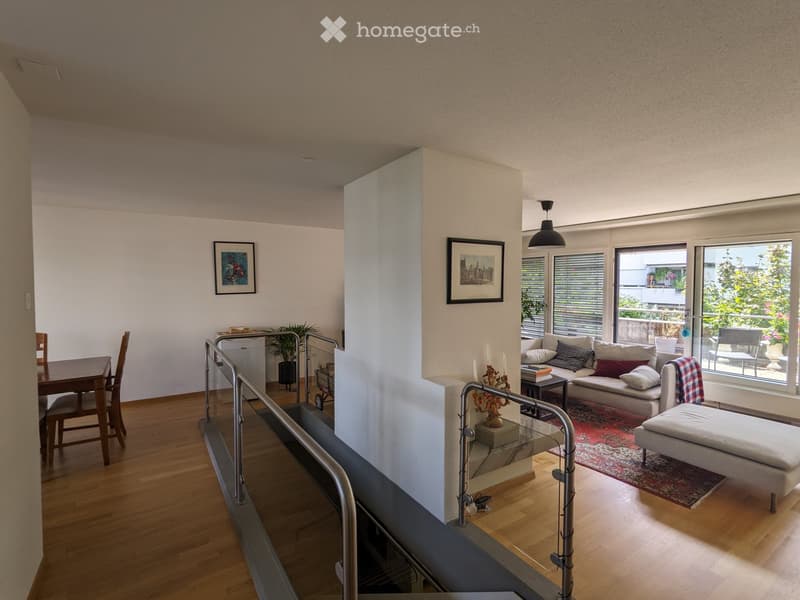 Open-plan living and dining area with wooden flooring, large windows, a sofa, ottoman, dining table, chairs, candle holder, picture frames, and a plant.