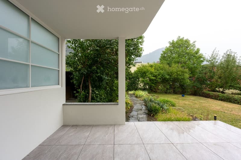 A modern outdoor patio with tiled flooring, a raised section overlooking a landscaped garden with trees, shrubs, and a grassy lawn.