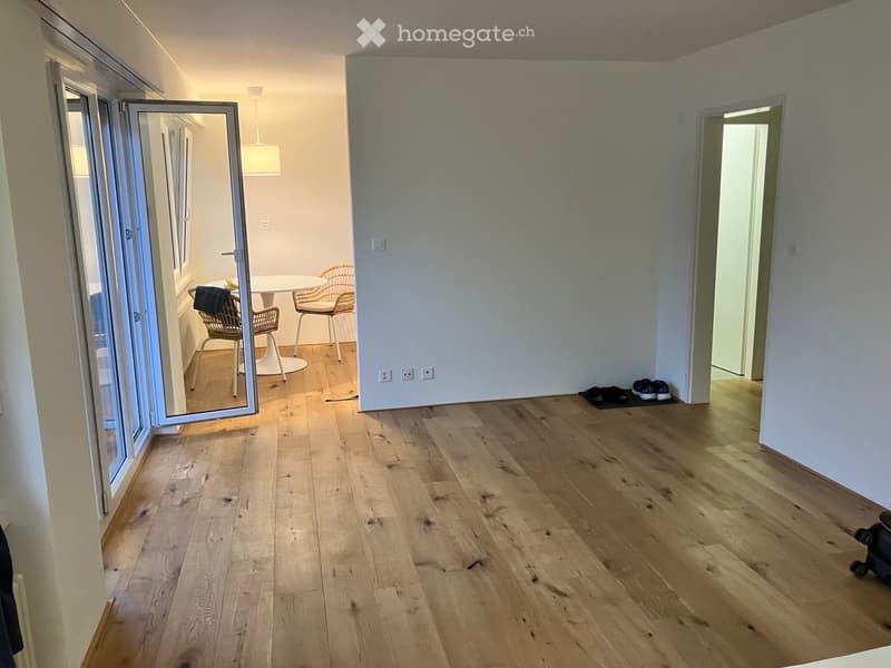Empty hallway with wooden flooring, white walls, and a large open door leading to another room. A small table and chairs are visible in the adjacent room.