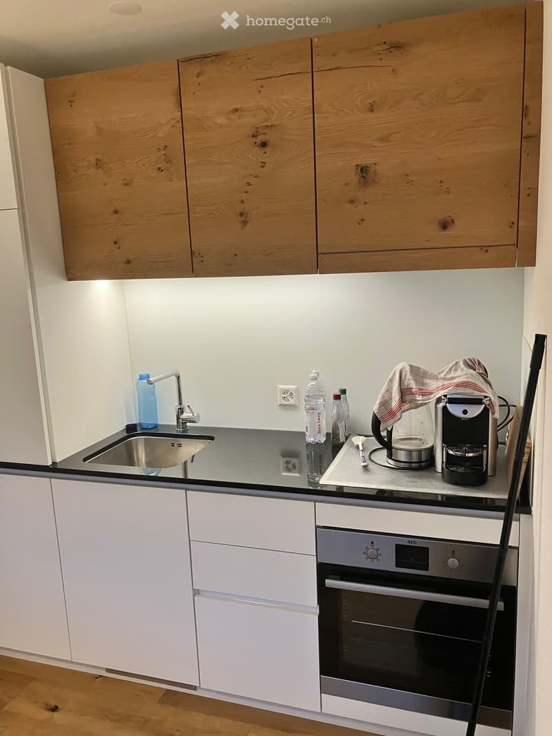 Modern kitchen with white cabinets, wooden upper cabinets, stainless steel sink, oven, coffee maker, kettle, and various bottles on the counter.