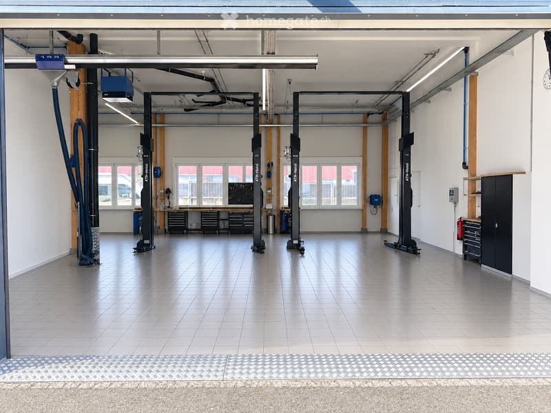 A spacious, well-lit garage with multiple hydraulic lifts, tool cabinets, workbenches, and storage units. The floor is tiled, and the walls are white with large windows allowing natural light.