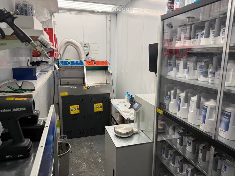 A well-organized workshop with a spray gun on the left, shelves filled with containers, a machine in the center, and a table with various tools and equipment.