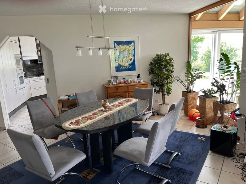 Furnished Room. Dining area with a round table, six chairs, and a kitchen area with white cabinets, microwave, and oven. There are potted plants, a framed picture, and a small table with decorative items.