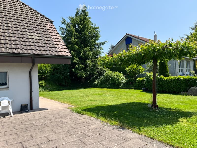 Garden / Outdoor Area. Well-maintained garden with a paved pathway, a white plastic chair, and a tree providing shade. The lawn is neatly trimmed, and the house has a tiled roof and white exterior.