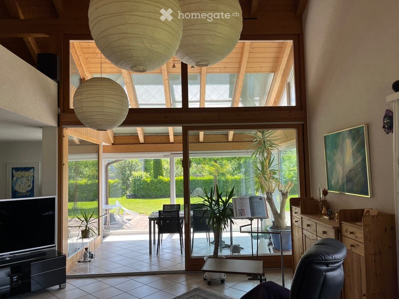 Furnished Room. Bright living room with large windows and sliding glass doors leading to a patio. Features a sofa, coffee table, chairs, a TV, potted plants, and a wooden cabinet.