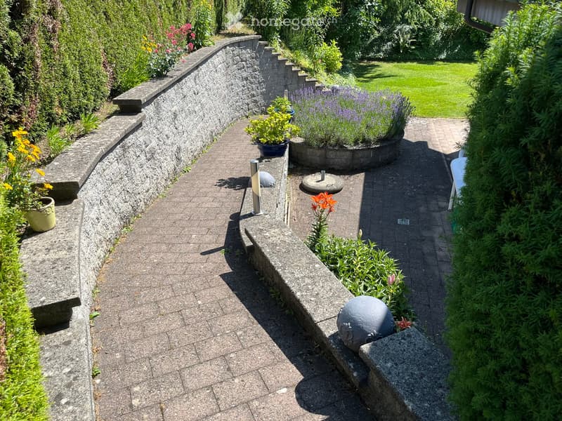 Garden / Outdoor Area. A curved garden pathway lined with stone walls, potted plants, and decorative spheres, leading to a grassy lawn.
