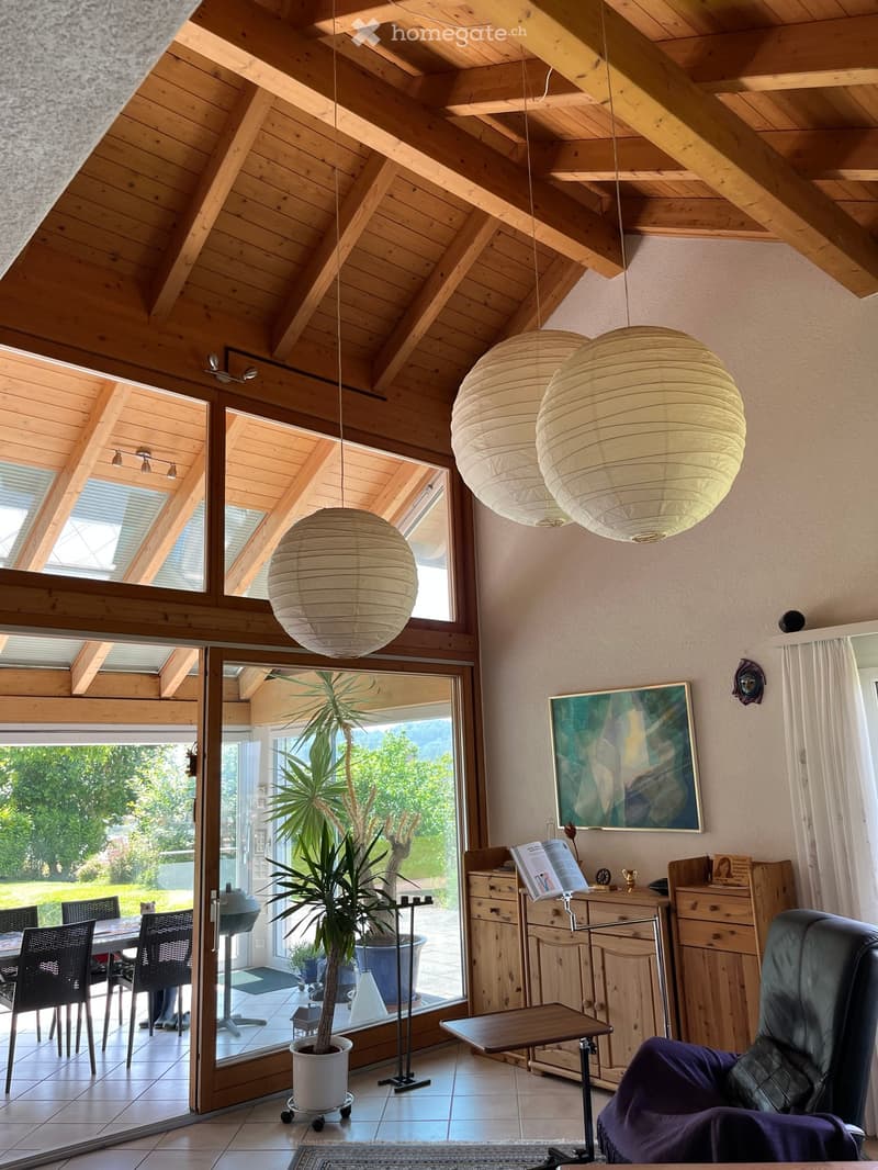 Furnished Room. Bright living space with wooden beams, large windows, hanging paper lanterns, a leather chair, a small table, a potted plant, and a painting on the wall.