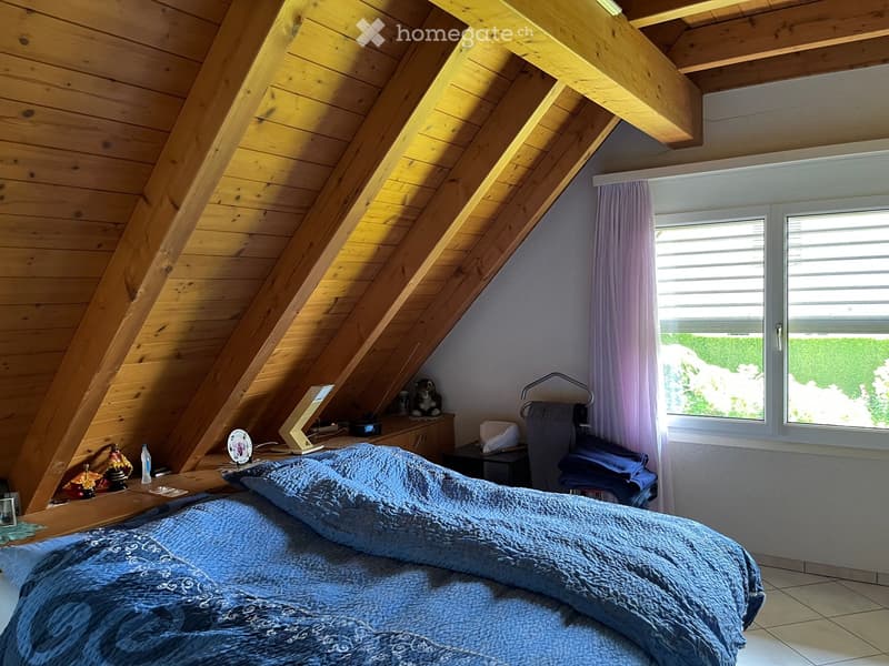 Bedroom. Cozy attic bedroom with wooden slanted ceilings, large windows with purple curtains, a neatly made bed with blue bedding, a nightstand with a table lamp, and decorative items on the windowsill.