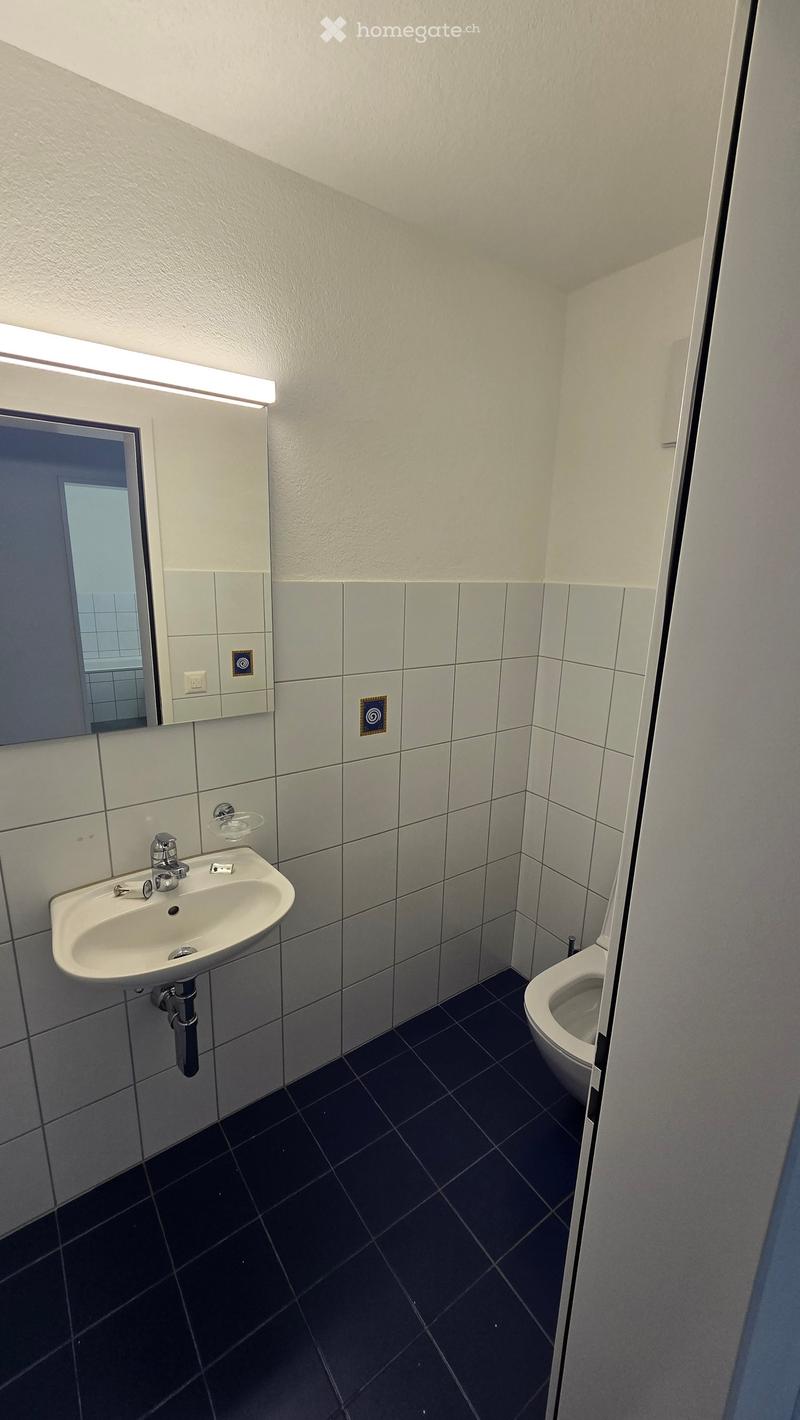 Modern bathroom with white tiled walls, dark tiled floor, a white sink with chrome faucet, and a toilet. A mirror with built-in lighting is mounted above the sink.