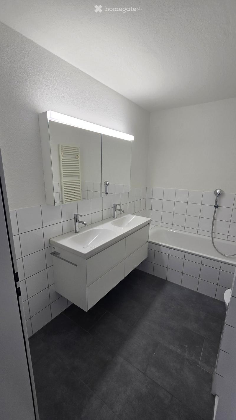 Modern bathroom with white tiles, a double sink, a large mirror with built-in lighting, and a bathtub.