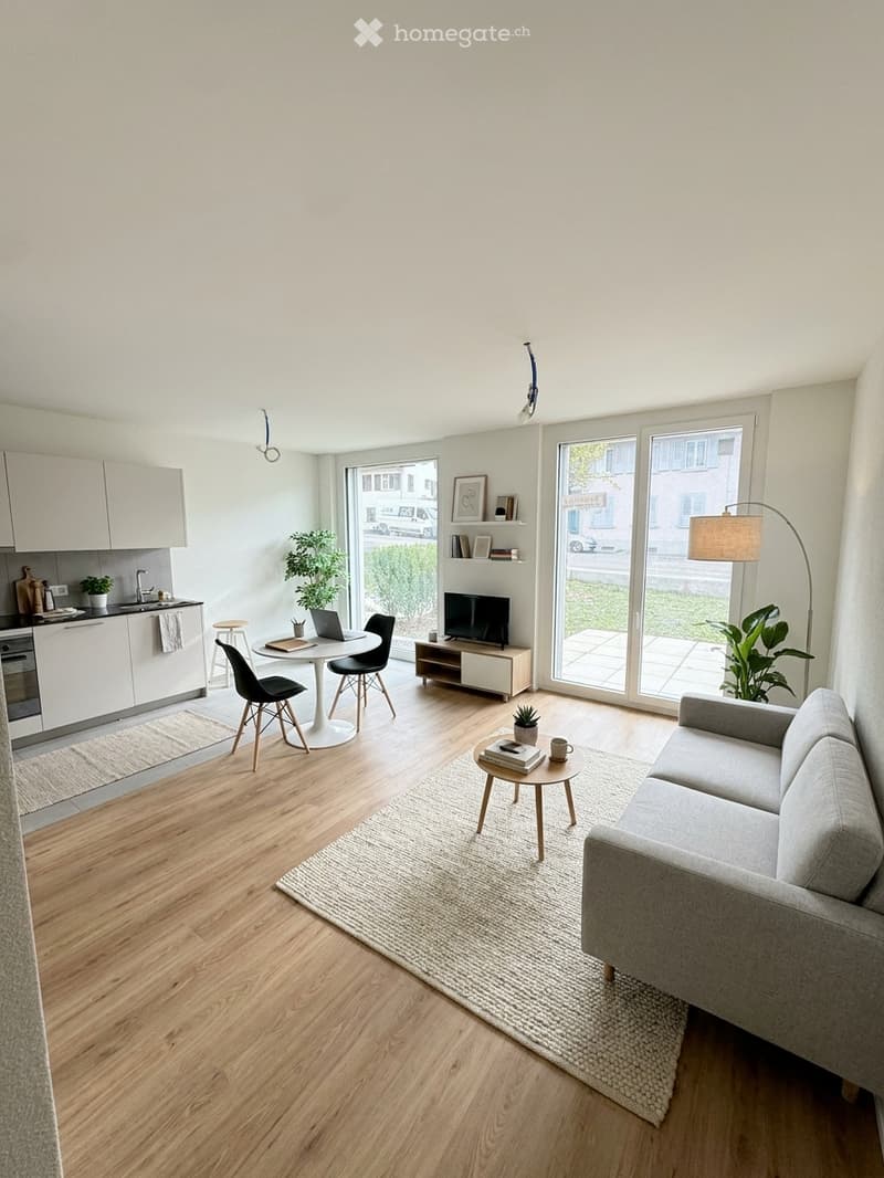 Modern open-plan living and dining area with light wood flooring, white walls, and large windows. Features a grey sofa, coffee table, dining table with chairs, a laptop, TV, floor lamp, and kitchen area with cabinets, oven, and sink.