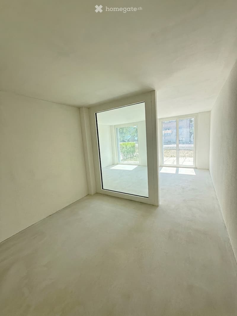 Empty room with light-colored walls and floor, featuring large windows and sliding glass doors leading to an outdoor area.